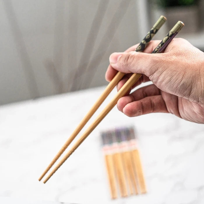 Hand holding a pair of wooden chopsticks with a blurred pack of chopsticks in the background.