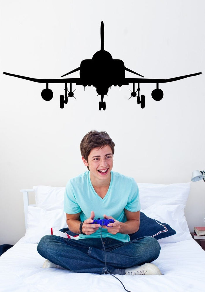 A young man sitting cross-legged on a white bed holding a purple video game controller, with a large black airplane silhouette decal on the wall above his head.
