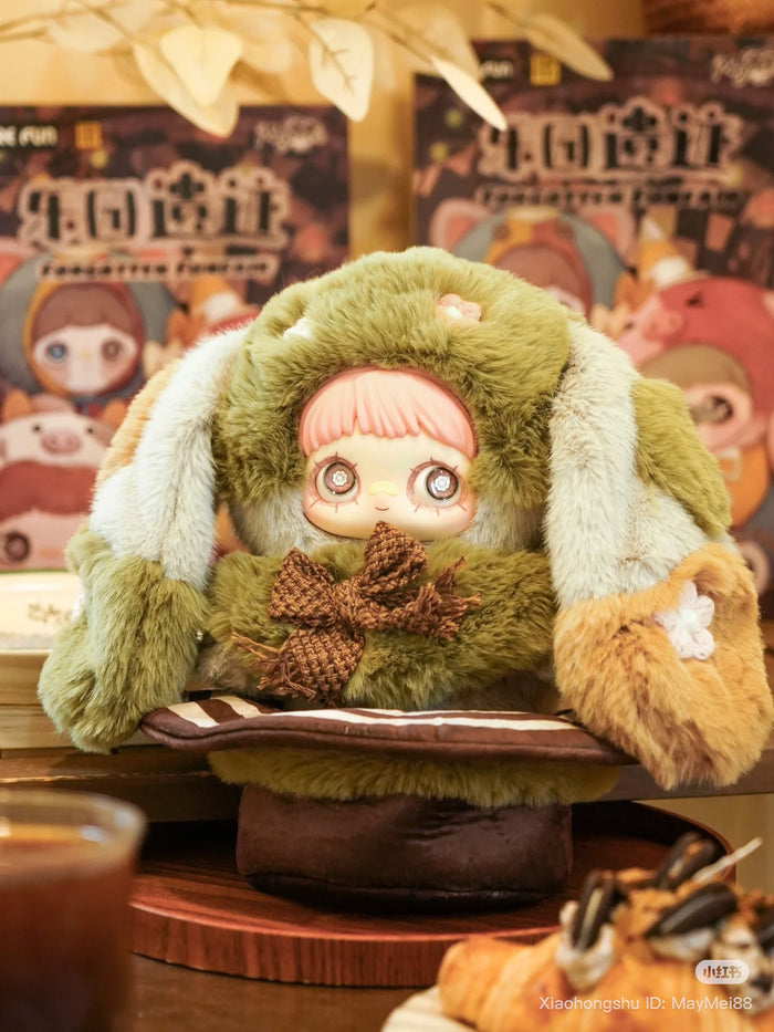 Maymei 400% Forgotten Funfair Plush Keychain Blind Box figure in green bunny suit, seated with coffee and pastry, colorful packaging blurred behind.
