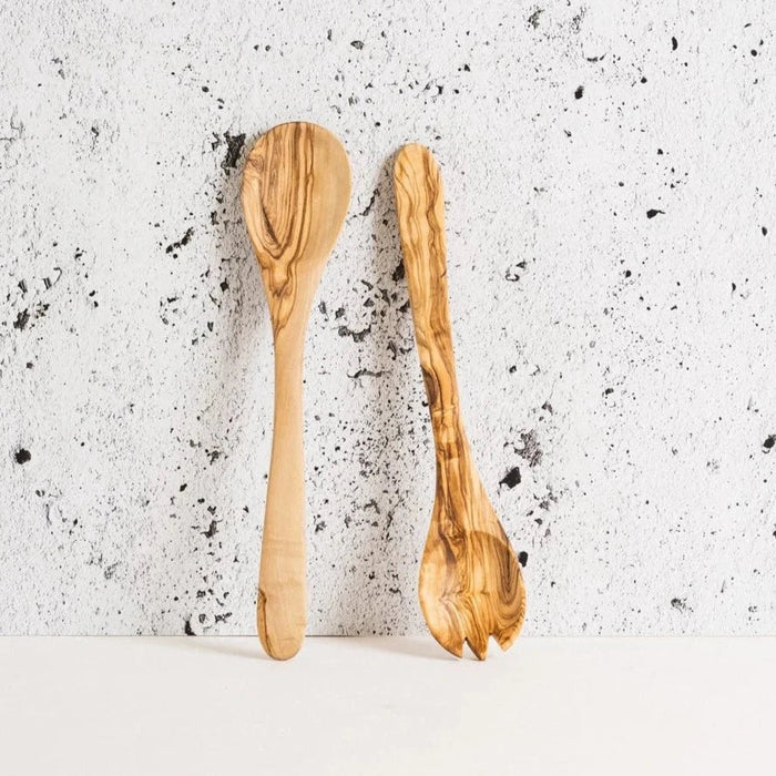 A set of olive wood utensils, including a spoon and a spork, displayed against a white background with a textured wall.
